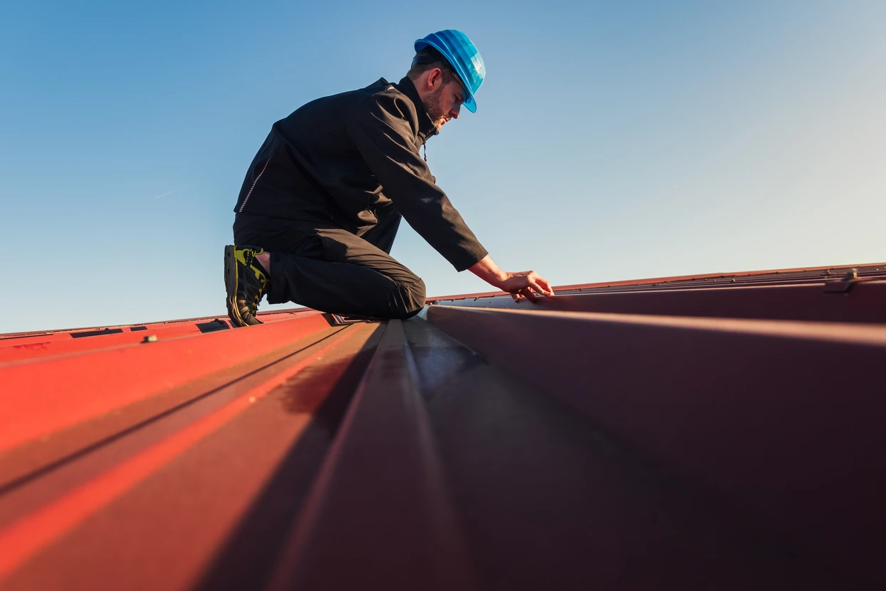 Ouvrier couvreur travaillant sur une toiture en tuiles rouges, portant casque de protection bleu et harnais de sécurité
